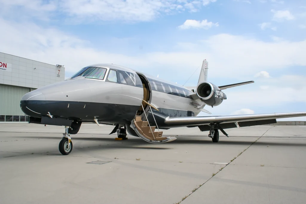 This image shows a Cessna Citation XLS, managed by Avcon Jet North America, parked on the tarmac. The jet is captured from a front-side angle with its boarding stairs deployed, emphasizing its streamlined design and polished exterior. A clear sky and an airport building in the background complete the setting.