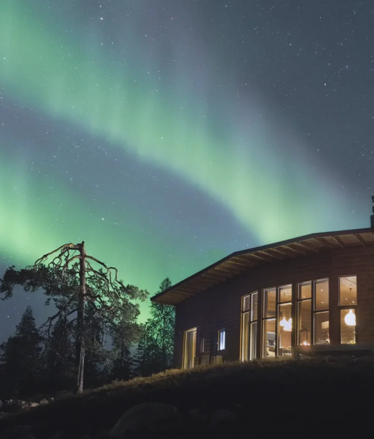 Octola Villa illuminated under the mesmerizing Northern Lights, surrounded by a starry sky and tranquil forest, offering a magical winter escape in the Arctic wilderness.