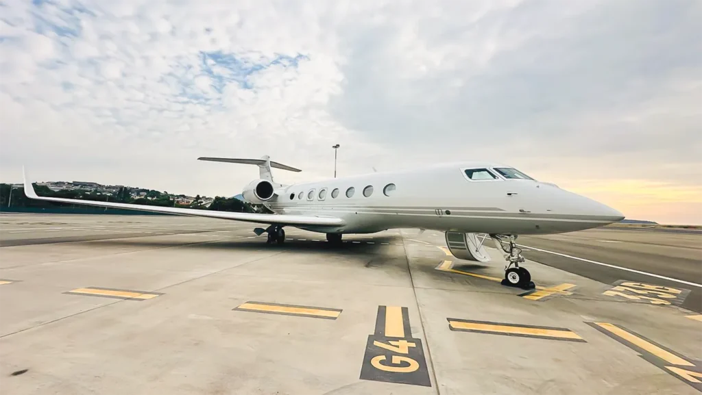 Featured aircraft from Avcon Jet: a Gulfstream G650ER private jet with a sleek white design, parked on a sunlit tarmac with a scenic backdrop of hills and a pastel-colored sky.